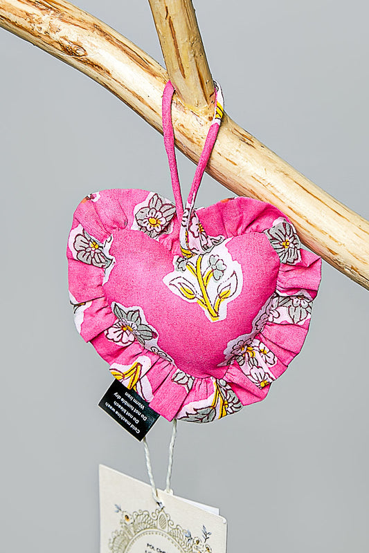 Pink heart-shaped fabric bag charm with floral design hanging on a branch against a gray background