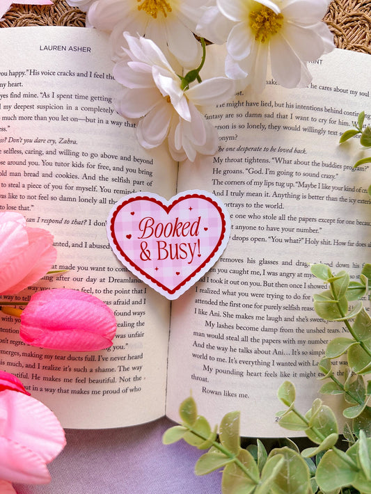 Heart-shaped 'Booked & Busy!' Sticker on an open book with flowers and leaves.