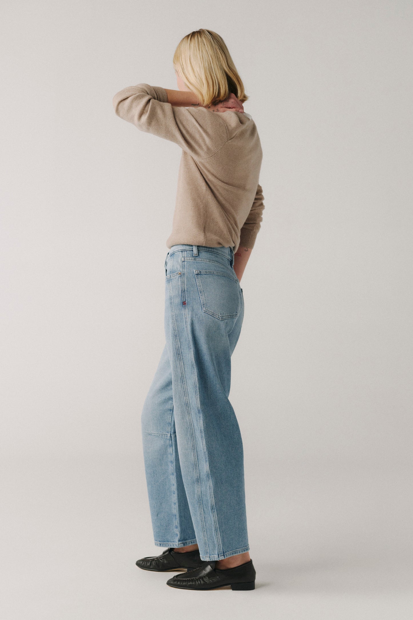 Person wearing a beige sweater and light blue jeans standing against a plain background