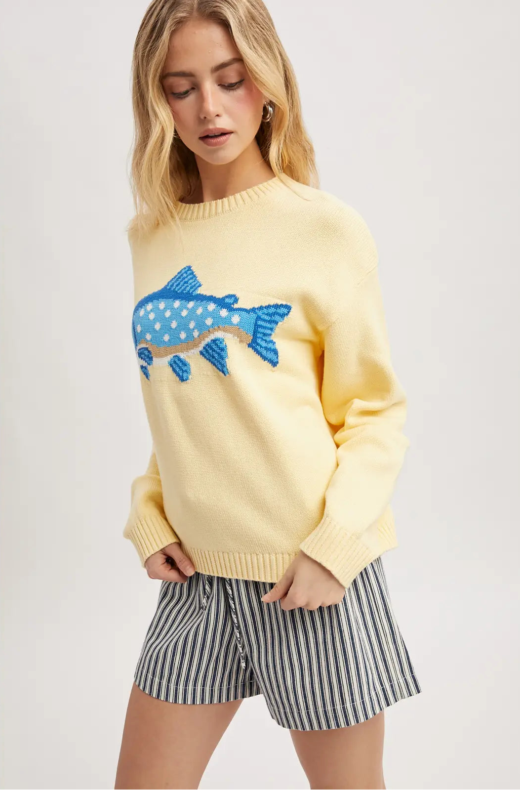 Woman wearing a yellow sweater with a blue fish design and striped shorts on a light gray background