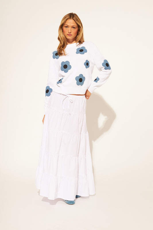 Woman wearing a white sweater with blue floral patterns and a white skirt on a light background
