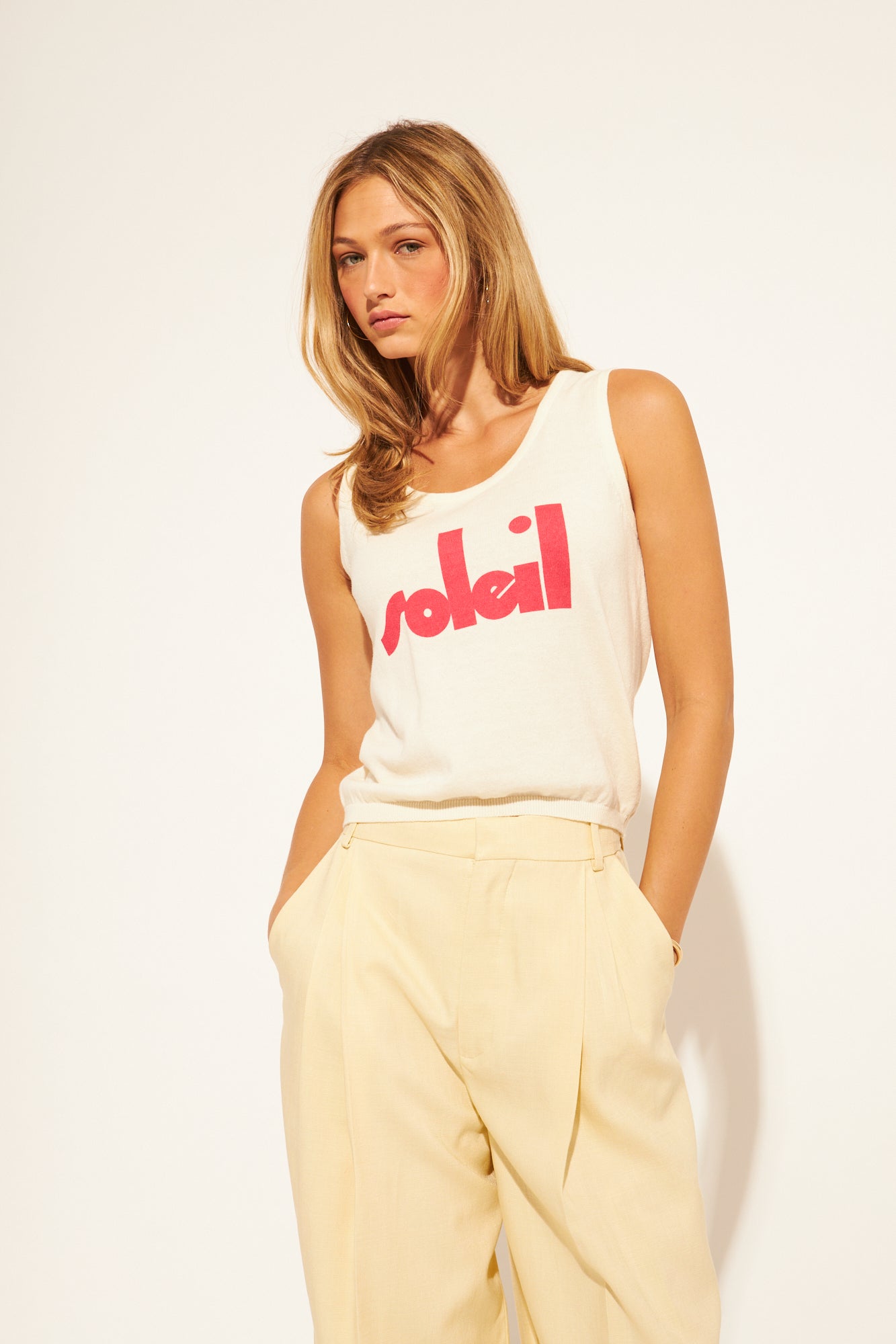Woman wearing a white tank top with red text and beige pants on a white background