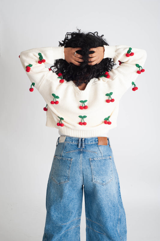 Person wearing a white sweater with red cherry patterns and blue jeans on a light gray background