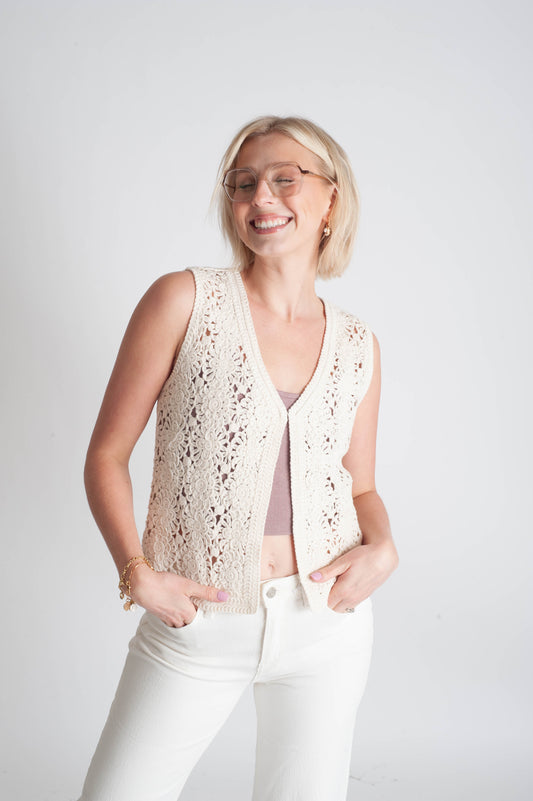 Woman wearing a beige lace vest over a pink top and white pants on a light gray background