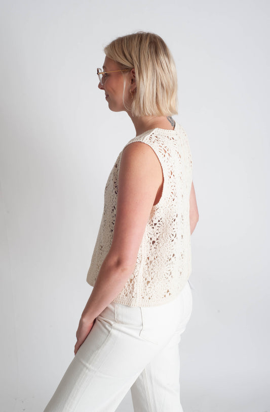 Woman wearing a sleeveless lace top and white pants against a plain background