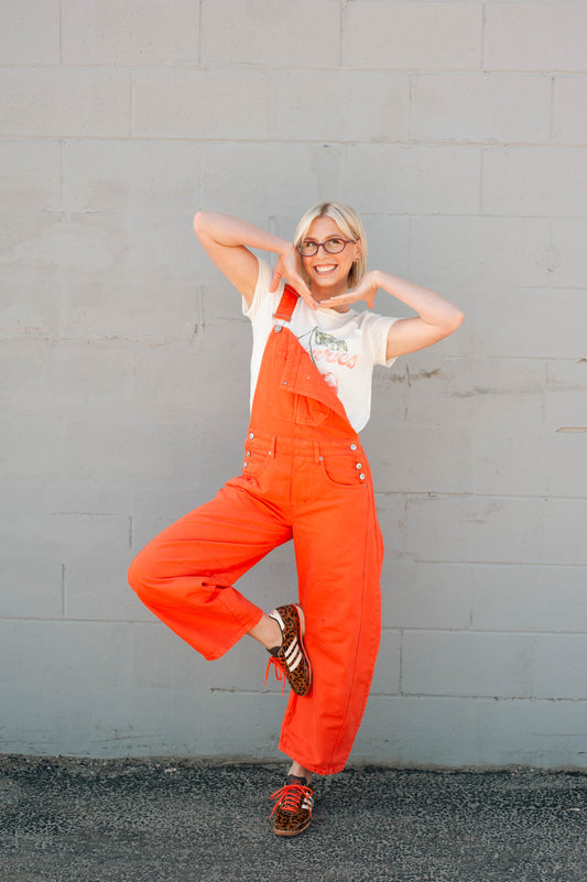 Person wearing bright orange overalls and a white t-shirt against a gray wall.