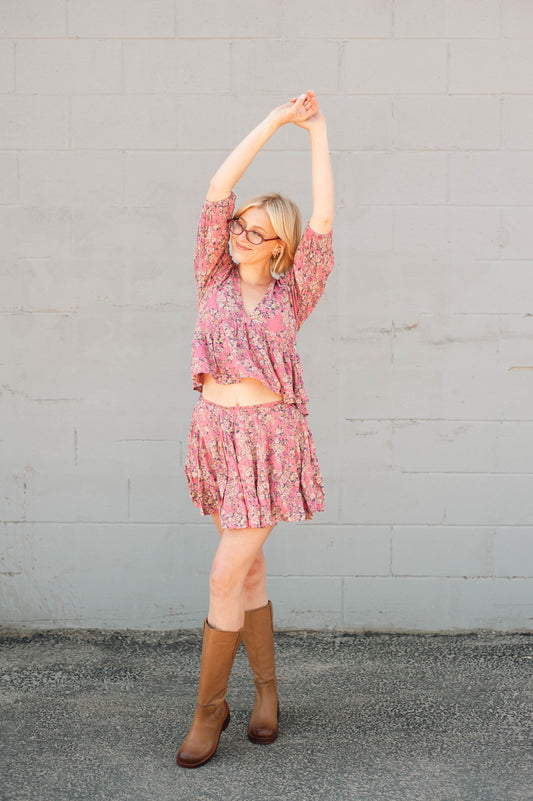 Woman in a pink floral dress and brown boots stretching against a gray wall.