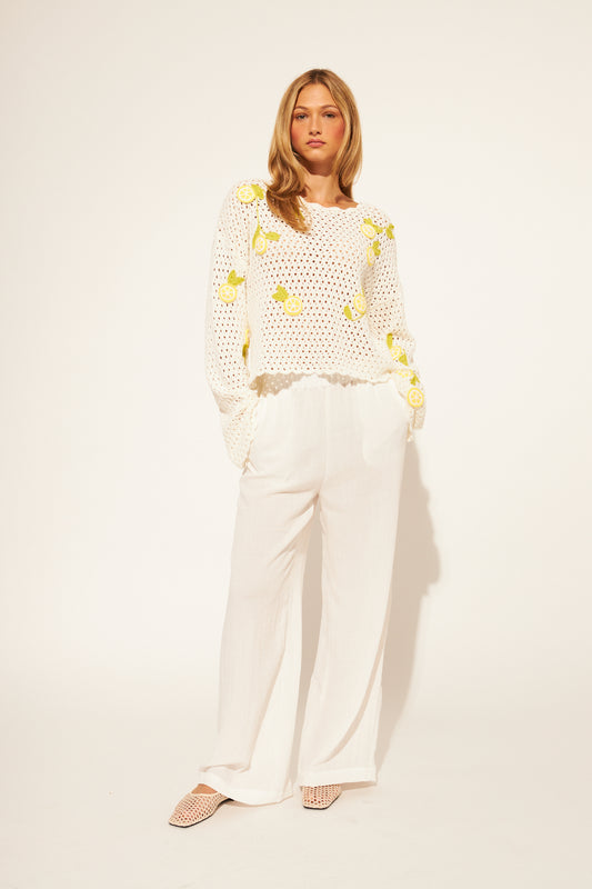 Woman wearing a white crochet top with yellow floral patterns and white pants on a plain background