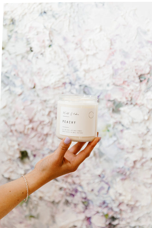 Hand holding a candle labeled 'Peachy' against a floral background