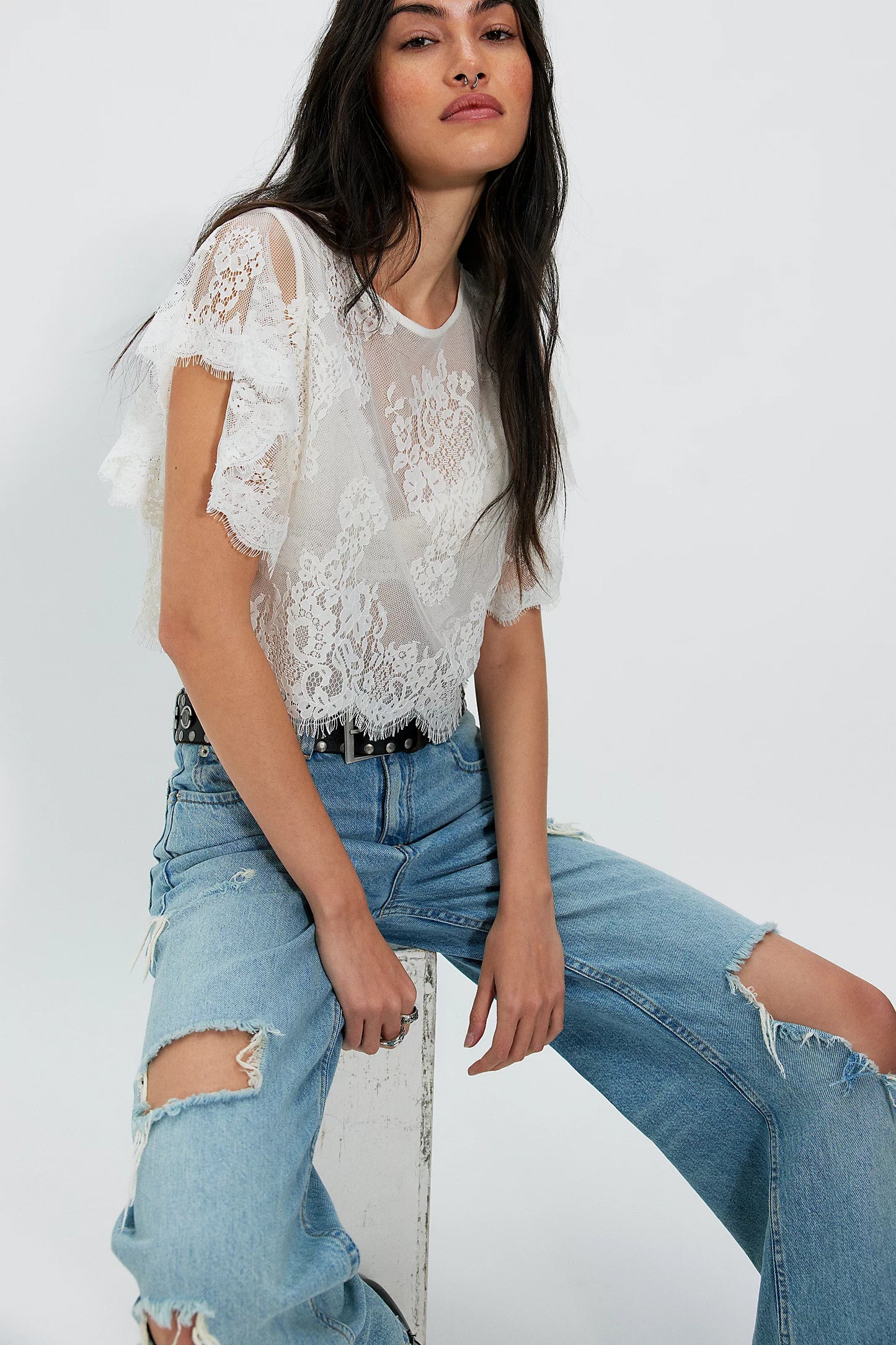 Woman wearing a lace top and ripped jeans sitting on a white stool against a white background
