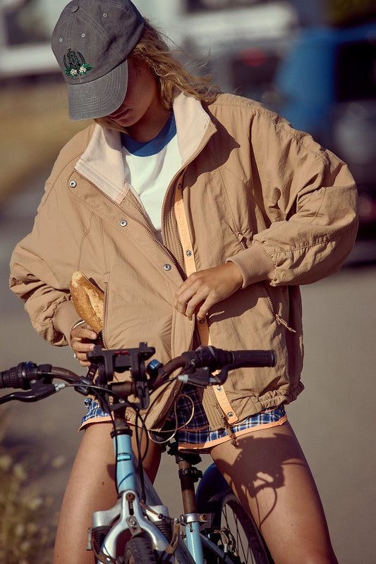 Person on a bicycle wearing a beige jacket and plaid shorts, holding a loaf of bread.