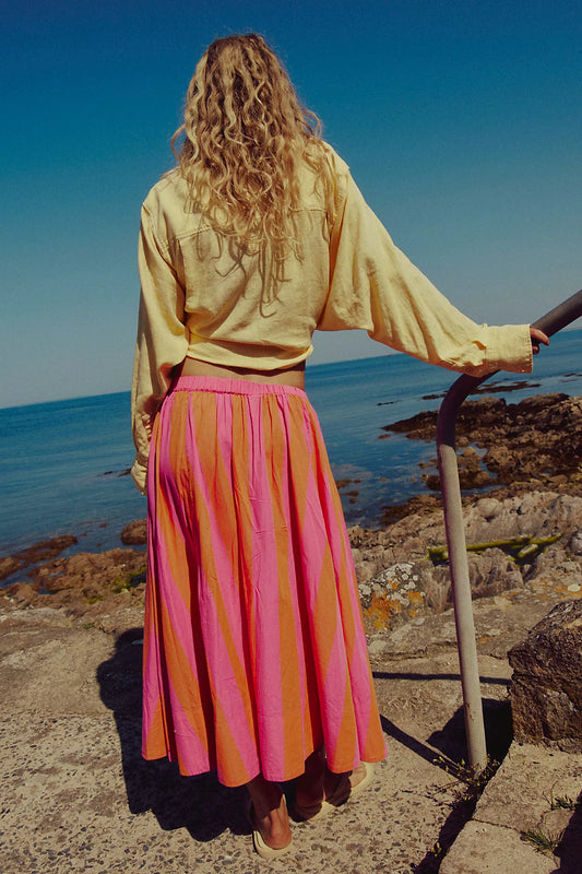 Person wearing a yellow top and pink skirt standing on a rocky coastal path.