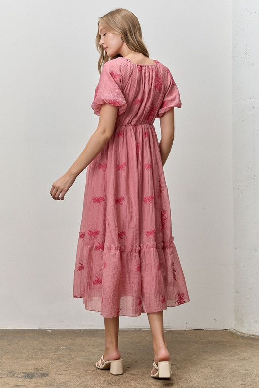 Woman wearing a pink dress with butterfly patterns against a white wall.