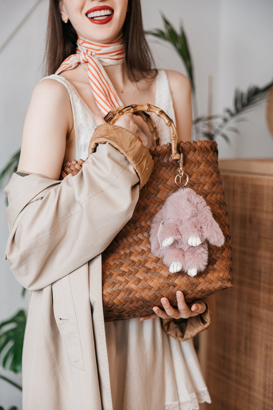 Woman holding a woven handbag with a plush bunny toy attached, wearing a beige coat and striped scarf.