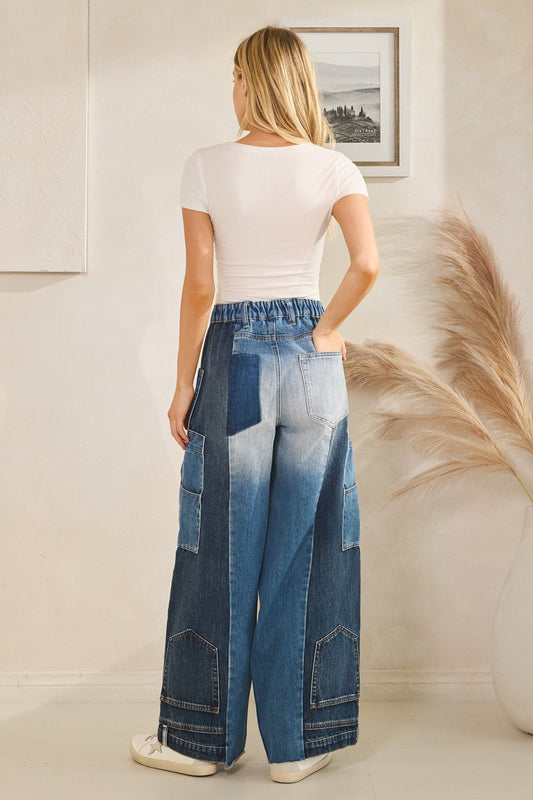 Person wearing a white t-shirt and blue patchwork wide-leg jeans in a minimal indoor setting.