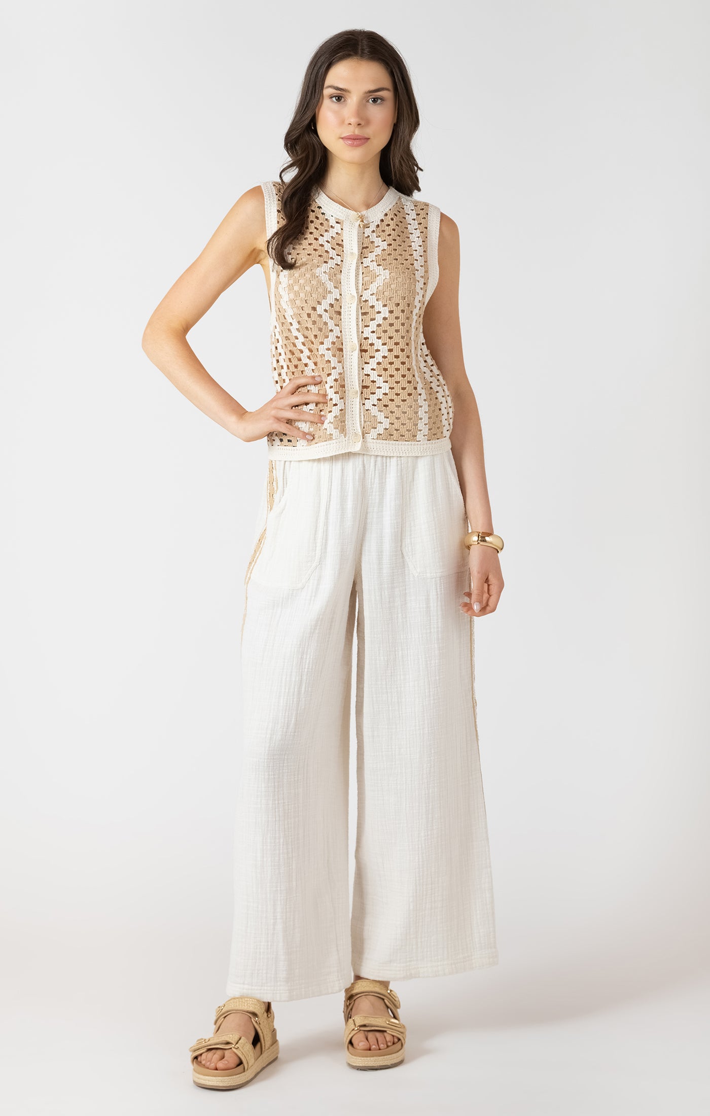 Woman wearing a sleeveless beige top with a mesh design and white pants on a white background