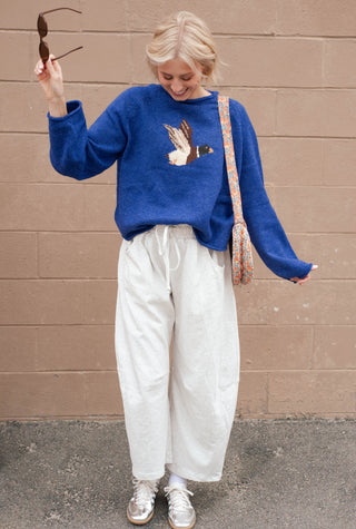 women wearing a duck sweater and grey sweatpants against a brick wall.