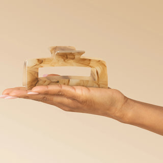 A hand holding a large tortoiseshell hair claw clip against a solid beige background.