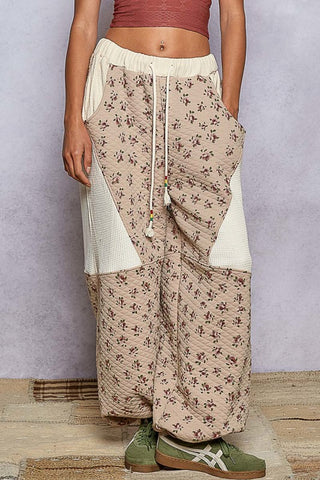 Person wearing a floral patterned pants with a plain white top and green shoes on a wooden floor.