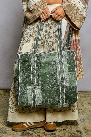 A person wearing a patchwork floral dress and brown shoes holds a large green and white patchwork bandana-print tote bag with striped handles, standing on a patterned rug against a gray wall.