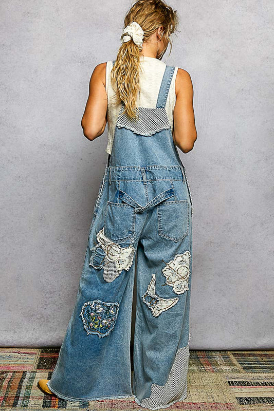 Person wearing denim overalls with floral patches against a plain wall.