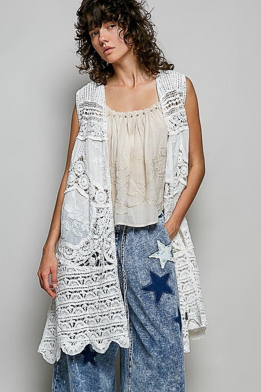 Woman wearing a white lace vest over a beige top and blue jeans with star design on a gray background