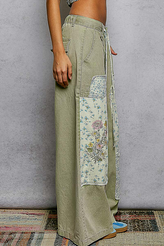 Person wearing olive green pants with floral patch and fringe details against a gray wall.