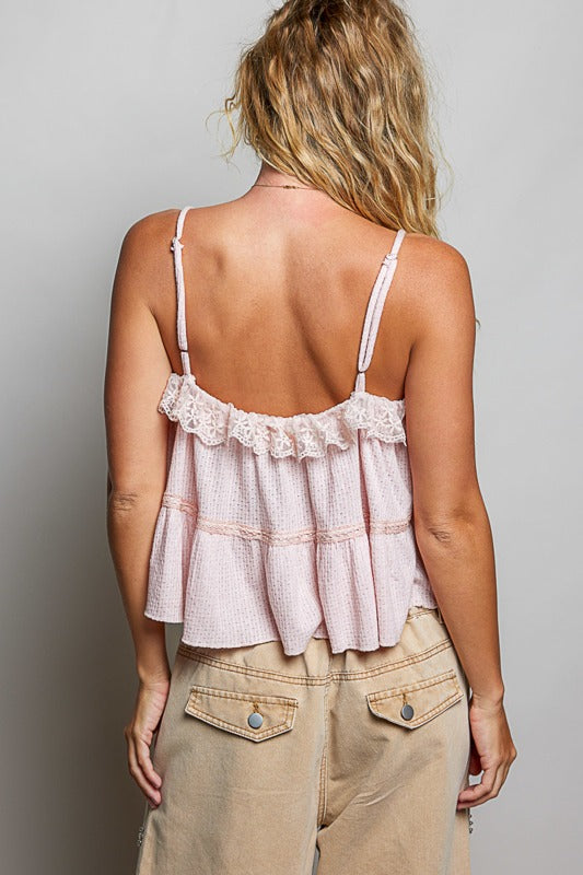 Woman wearing a light pink lace-trimmed top and beige pants against a gray background