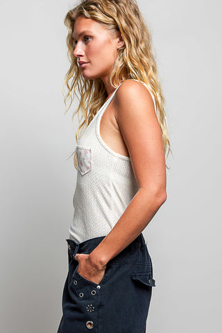 A woman in profile wearing a white textured tank top with a chest pocket and dark navy pants, photographed against a light gray studio background.
