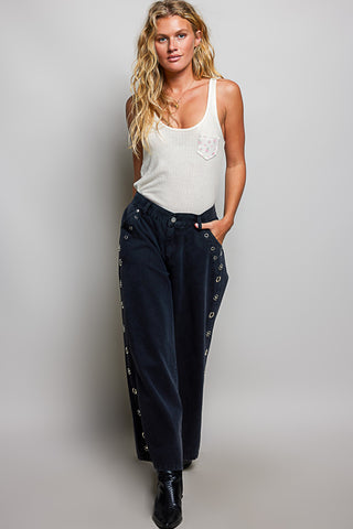 A woman modeling a pair of dark navy wide-leg jeans with silver grommets down the sides, paired with a white tank top and black boots, against a light gray studio background.