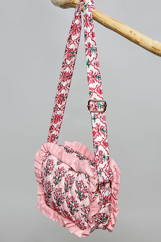 A pink ruffled shoulder bag with a floral print and adjustable shoulder strap, hanging from a wooden dowel against a solid gray background.