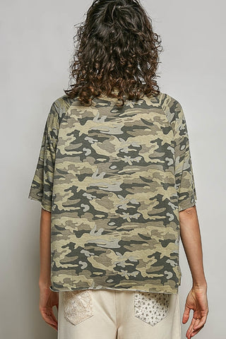 A person shown from the back wearing an oversized short-sleeve t-shirt with a green, beige, and brown camouflage pattern, paired with cream-colored pants.