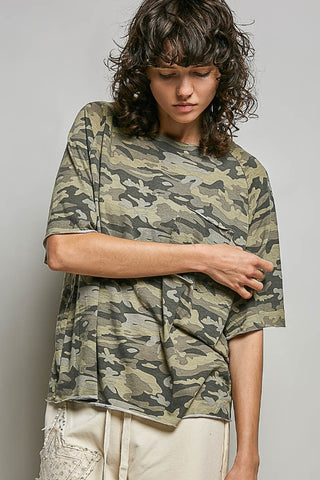A model wearing an oversized short-sleeve camouflage t-shirt in olive green, beige, and gray tones, paired with cream-colored pants, against a neutral gray studio background.