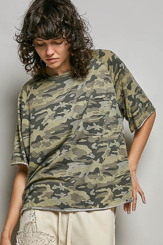 A woman wearing an oversized short-sleeve camouflage T-shirt with a chest pocket, paired with light beige pants, photographed against a plain light gray studio background.