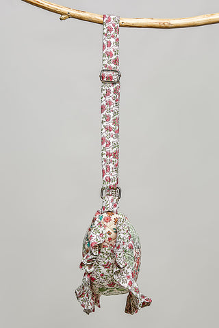 A fabric pet sling bag with a floral print, hanging from a natural branch. The bag features a long strap with metal buckles and a ruffled bottom edge.