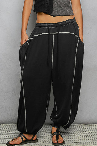 Black harem-style pants with white contrast stitching and a drawstring waist, shown on a model from the waist down against a gray textured wall.