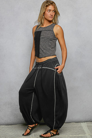 A woman modeling a two-piece outfit consisting of a gray sleeveless crop top with a black side panel and black harem pants with white piping details, standing against a textured gray wall.