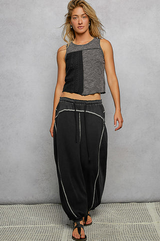 A woman modeling a sleeveless patchwork crop top in heather gray and black, paired with loose black drawstring pants featuring white piping details, set against a textured gray studio backdrop.
