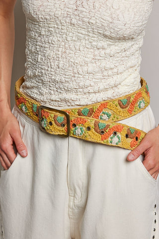 A close-up of a person wearing a wide, heavily embroidered yellow belt with a brass buckle, paired with a white textured top and white pants.