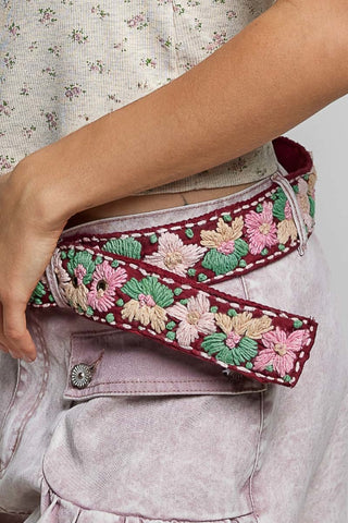 A close-up of a person wearing a wide embroidered belt with pink and green floral patterns over a dusty pink denim skirt.
