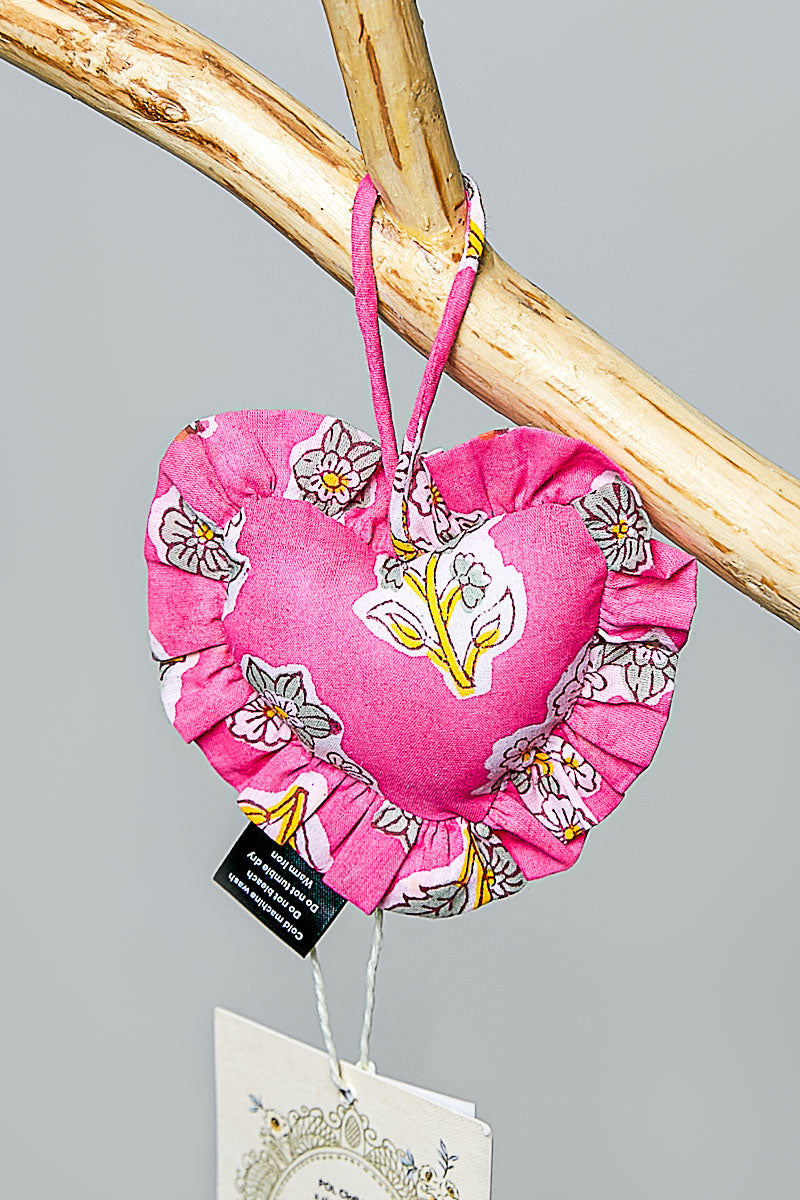Pink heart-shaped fabric bag charm with floral design hanging on a branch against a gray background
