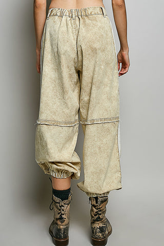 A person shown from the back wearing loose, oversized cropped pants in a light beige acid-wash or mineral-wash fabric, paired with dark lace-up boots against a plain light gray studio background.