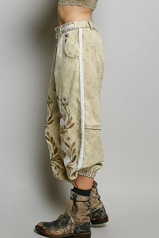 A person shown from the waist down wearing loose, cropped beige pants with a white side stripe, floral embroidery near the lower leg, and distressed brown boots against a plain light gray studio background.