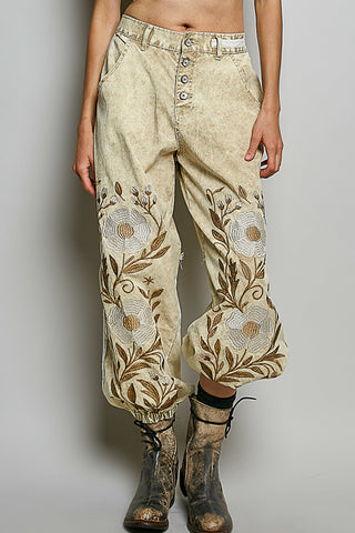 A person wearing loose, high-waisted beige pants with large embroidered floral motifs in tan and brown, paired with dark lace-up ankle boots, against a plain light gray studio background.