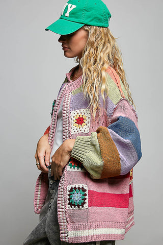 A woman wearing a colorful patchwork knit cardigan with crochet-style squares, a green baseball cap, and gray jeans, photographed against a plain light gray studio background.