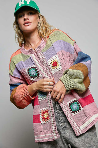 A woman wearing a colorful striped crochet cardigan with granny square patches, a green baseball cap with 'NY' on it, and gray jeans, photographed against a plain gray studio background.