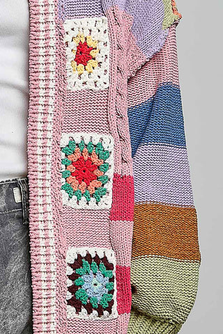 A close-up of a chunky knit cardigan featuring pink and pastel striped sections with colorful crocheted granny square patches, worn over a white top and gray jeans.