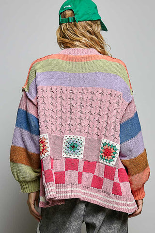 A person shown from the back wearing a colorful striped and patchwork knit cardigan, a green baseball cap, and gray pants against a plain light gray studio background.