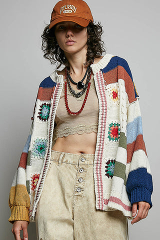 A model wearing a colorful crochet cardigan with granny square patterns, paired with a beige crop top, beige jeans, layered necklaces, and an orange cap, set against a plain gray studio background.