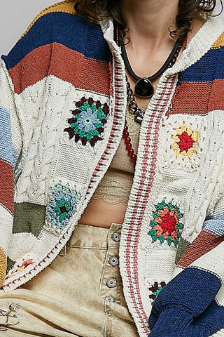 A close-up of a person wearing a chunky knit cardigan with colorful striped panels and crocheted floral motifs. The cardigan features cream, navy, rust, and light blue sections with embroidered flower details.